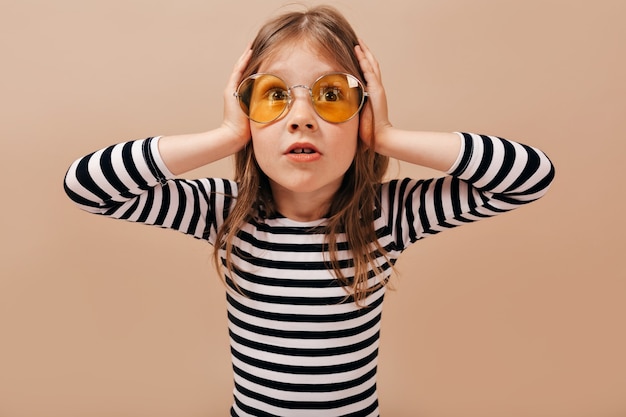 Free Photo adorable little cute girl wearing round yellow glasses looks worried, holding hands on the head and looking away