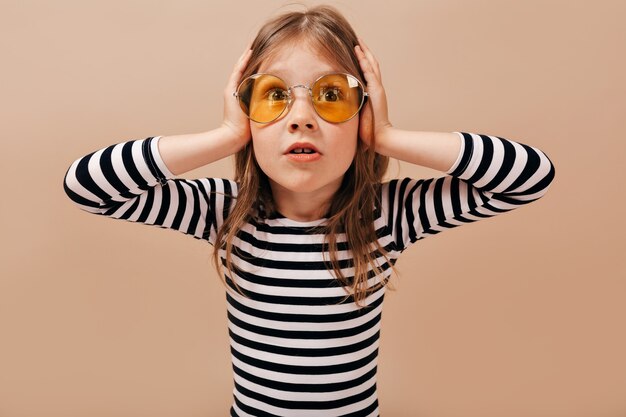 Adorable little cute girl wearing round yellow glasses looks worried, holding hands on the head and looking away