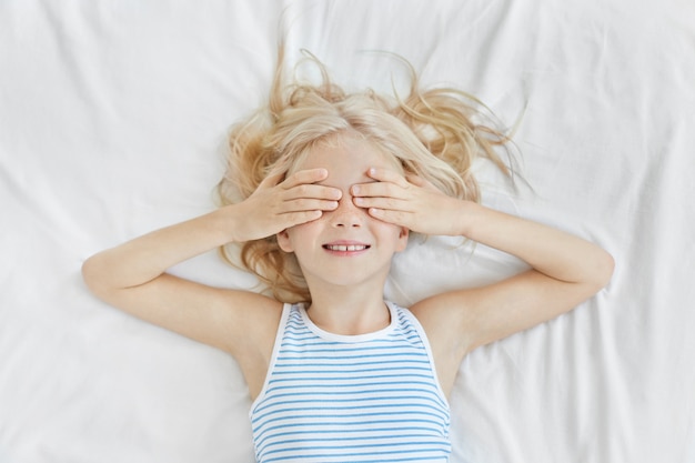 Free photo adorable little girl lying on white bedclothes, covering her eyes with hands, wearing sailor t-shirt, smiling before sleep. blonde kid with freckles having fun on bed not wanting to have sleep