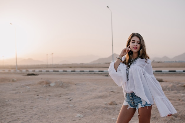 Free photo adorable long-haired woman in vintage blouse posing with eyes closed, touching her face with desert view. charming pretty young woman in white shirt dancing with pacific face expression