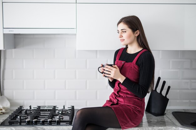 Adorable nice charming girl drinking coffee, sitting on table countertop in modern light white kitchen