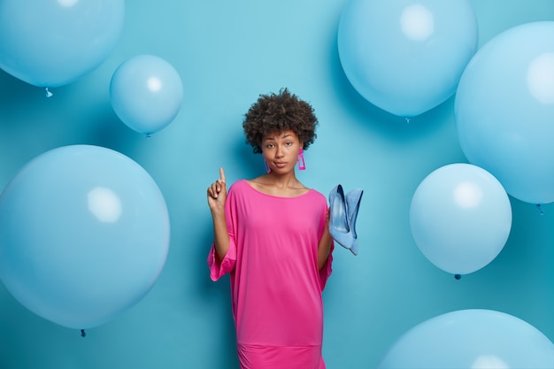 Free photo adorable serious dark skinned woman ready for upcoming date, points above and demonstrates store where she bought shoes, wears fashionable attire, stands against blue wall, inflated balloons