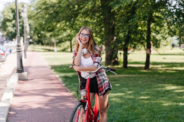 Adorable smiling girl posing in park with bicycle. Outdoor photo of relaxed lady posing on nature.