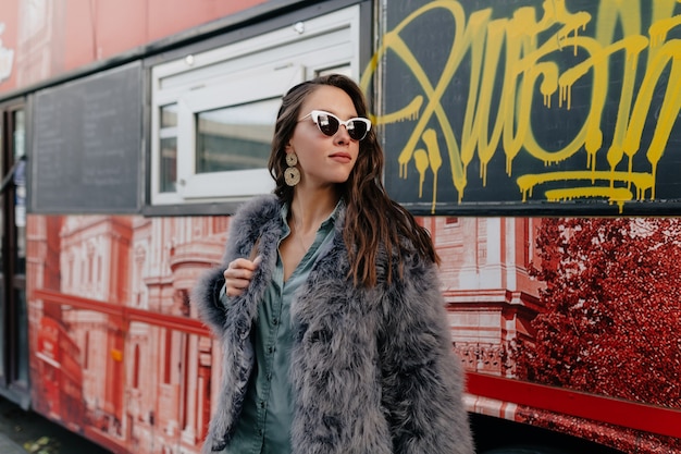 Adorable stylish lady with long wavy hair wearing fur coat posing during outdoor photoshoot.