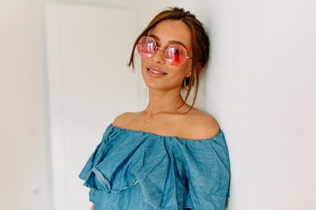 Adorable stylish tanned woman with dark hair wearing blue tshirt and pink round glasses posing at camera over isolated background