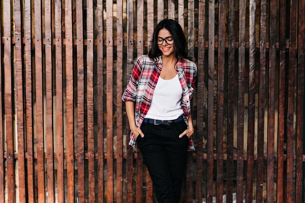 Adorable tanned female model enjoying spring photoshoot. Good-humoured caucasian girl in glasses chilling outdoor.