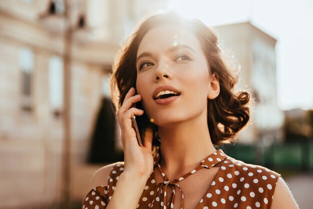 Adorable white woman calling someone while walking around town. Outdoor shot of dreamy caucasian girl standing on the street with cell.