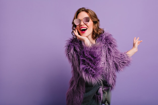 Adorable woman expressing true positive emotions during photoshoot in purple fur coat