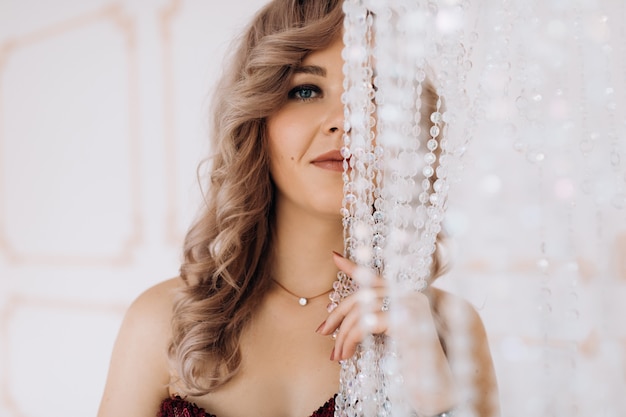 Free photo adorable woman in red burgundi dress poses in a bright luxury room with large chandelier