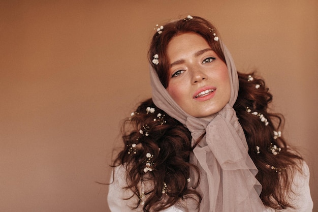 Adorable woman with freckles looks into camera with smile Lady in beige scarf on her head and with flowers in her red hair posing on beige background