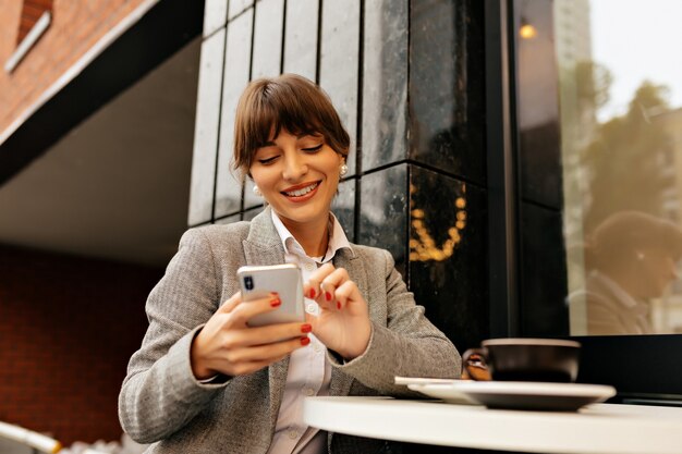 Adorable young brunette lady in grey jacket and white shirt using smartphone and works in laptop