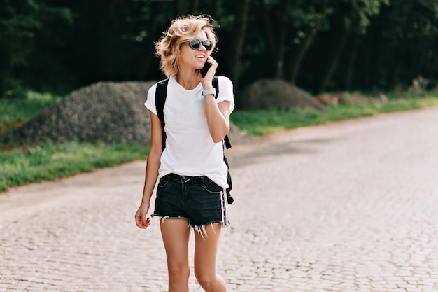 Free Photo adorable young charming lady with short hairstyle walking on the road with backpack and talking on the phone over mountains. travel mood, vacation, trip. wanderlust and travel concept.