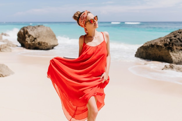 Adorable young woman in long red dress posing at wild beach with rocks. Outdoor shot of shapely girl walking near sea.