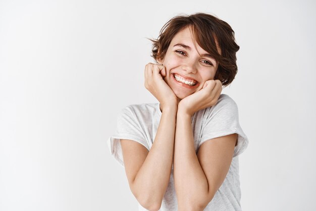 Adorable young woman smiling and looking silly, showing natural glowing skin without makeup, standing against white wall