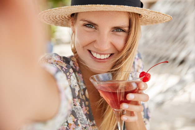 Free Photo adorable young woman with happy expression, holds fresh cherry cocktail, makes selfie, pose outdoor, has delighted look, glad to have summer rest in resort country. happiness and recreation concept