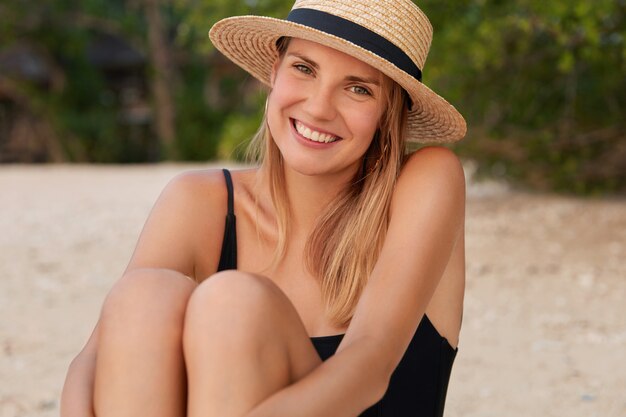 Adorable young woman with pleased expression, sunbathes on beach, sits on sandy beach, wears summer straw hat and black swimming suit, has tanned skin.