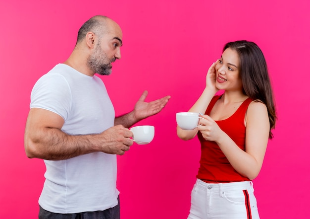 Adult couple both holding cups and looking at each other man telling something to woman and showing empty hand woman smiling and keeping hand on face 