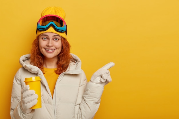 Adult female snowboarder with ginger hair, enjoys hot bevergae during winter time, wears ski clothing, points away on free space for your promotional content or text.