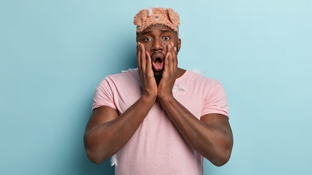 Adult man with dark skin, feels shocked after having not enough sleep, red eyes, wakes up too early, wears eyemask, casual t shirt, isolated over blue wall. Stunned Afro man in nightwear