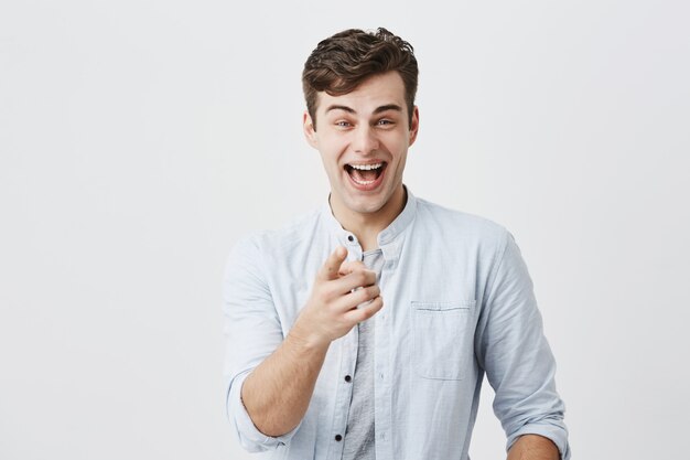 Advertising concept. Excited cheerful european man wearing light blue shirt raising eyebrows and exclaiming, pointing index fingers , choosing you, motivating and attracting customers.