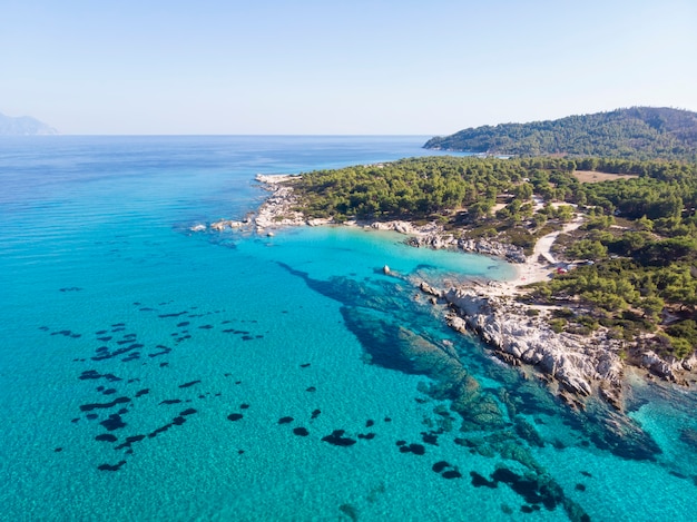 Aegean sea coast with blue transparent water, greenery around, rocks, bushes and trees, view from the drone, Greece