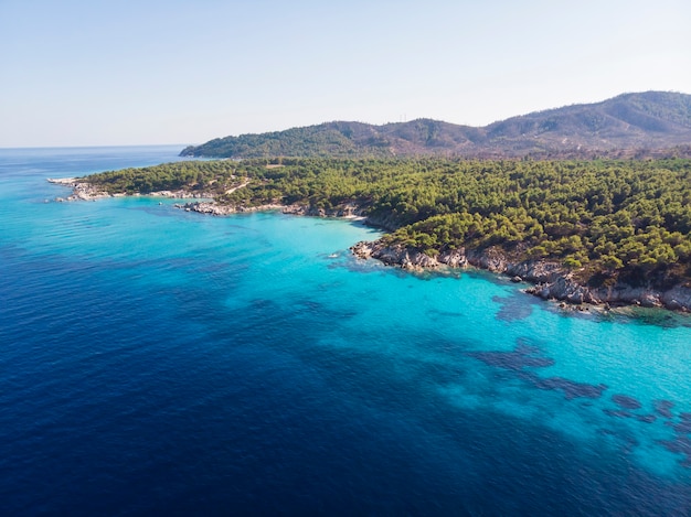 Aegean sea coast with blue transparent water, greenery around, rocks, bushes and trees, view from the drone, Greece