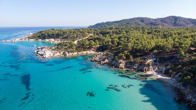 Aegean sea coast with blue transparent water, greenery around, rocks, bushes and trees, view from the drone, Greece