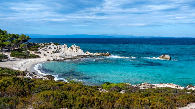 Aegean sea coast with greenery around, rocks, bushes and trees, blue water with waves, Greece