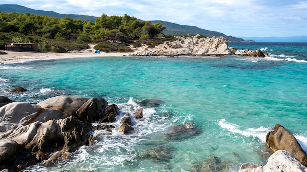 Aegean sea coast with greenery around, rocks, bushes and trees, blue water with waves, Greece