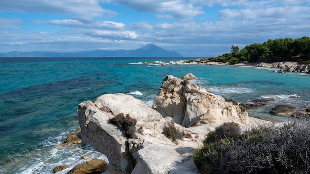 Aegean sea coast with greenery around, rocks, bushes and trees, blue water with waves, mountain