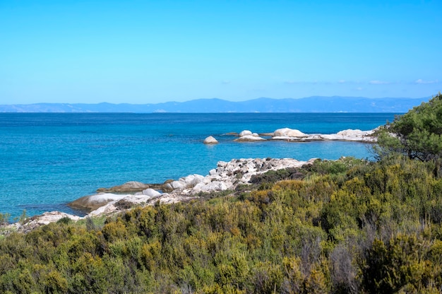 Aegean sea coast with rocks over the water and land in the distance, greenery on the foreground, blue water, Greece