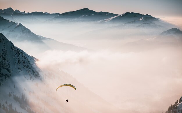 Aerial shot of a person parachuting down above the clouds near snowy mountains