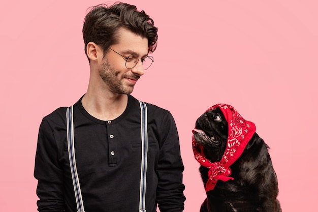 Free photo affectionate male dog owner looks with pleased expression and love at his funny pet with bandana on head, feels responsibility, play together at home, isolated over pink wall. pedigree puppy