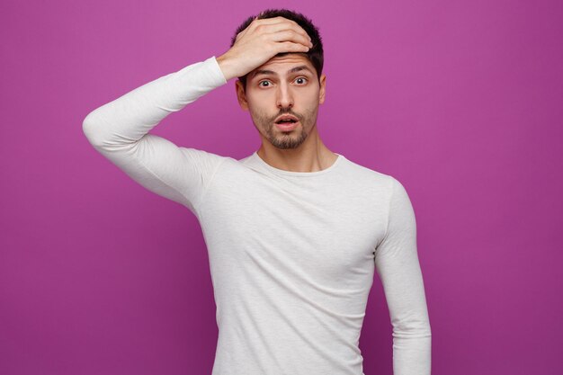 Afraid young handsome man keeping hand on forehead looking at camera on purple background