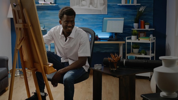 African american adult sitting in artwork studio drawing using pencil, vase design and white canvas on easel. Black young man with creative hobby making successful masterpiece and fine art