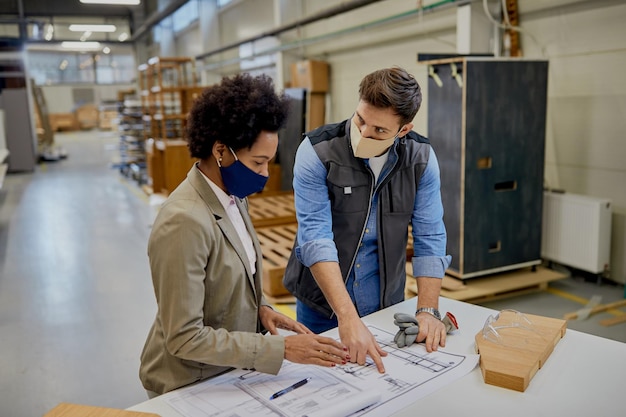 Free Photo african american businesswoman and carpenter wearing face masks while analyzing design plans at production facility