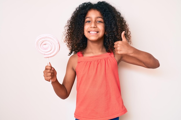 Free photo african american child with curly hair holding lollipop smiling happy and positive, thumb up doing excellent and approval sign