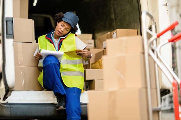 African American delivery woman talking on the phone while going through delivery list in a van