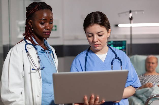 African american doctor health specialist and assistant using laptop, discussing talking, treatment and diagnosis, in hospital room with sick patient laying in bed with iv drip attached.