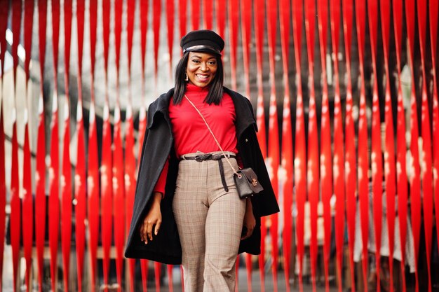 African american fashion girl in coat and newsboy cap posed at street against red background