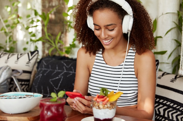 African American female with bushy hairstyle, listens online radio broadcast in headphones, connected to wireless internet in cafe, eats delicious dessert. People, technology, leisure concept