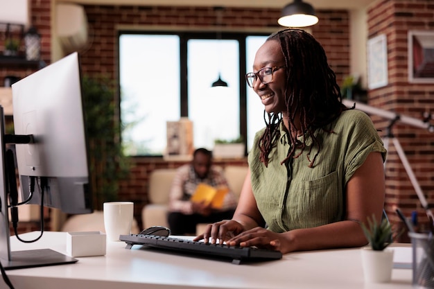 African american freelancer working remote typing and looking at computer screen while boyfriend is relaxing. Smiling programmer using pc to chat with friends while roommate is reading a book.