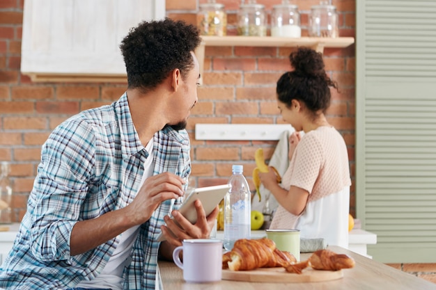 African American guy looks at wife or girfriend, asks her to give banana, sits at kitchen table, uses modern tablet computer