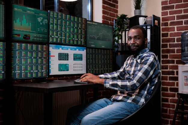 Free photo african american investment company trading agent sitting at workstation desk with multiple displays, looking at camera. financial broker in trading agency office with real time graphs on monitors.