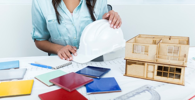 African-American lady on chair holding safety helmet near plan and model of house on table