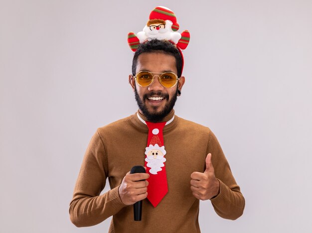 African american man in brown sweater and santa rim on head with funny red tie holding microphone smiling showing thumbs up standing over white background