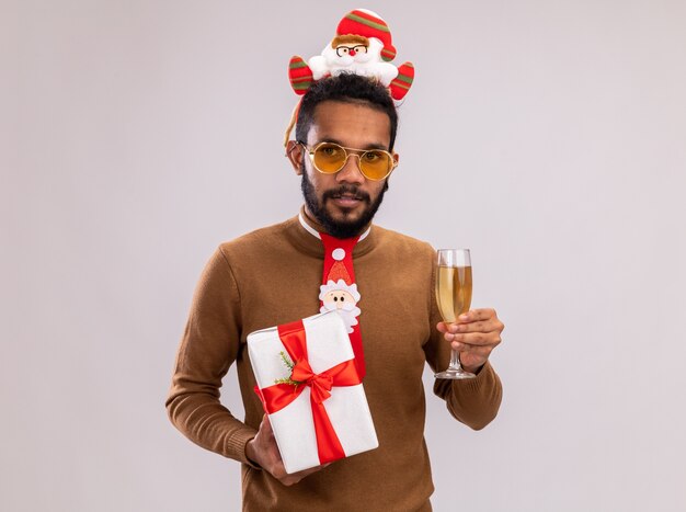 African american man in brown sweater and santa rim on head with funny red tie holding a present and glass of champagne looking at camera with confident expression standing over white background