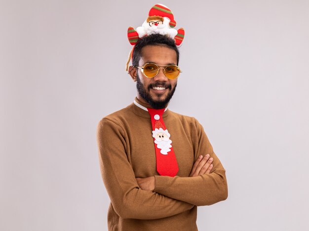 African american man in brown sweater and santa rim on head with funny red tie looking at camera smiling confident standing over white background