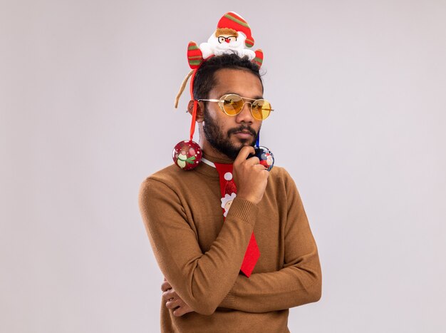 African american man in brown sweater and santa rim on head with funny red tie with christmas balls on his ears looking at camera with pensive expression standing over white background