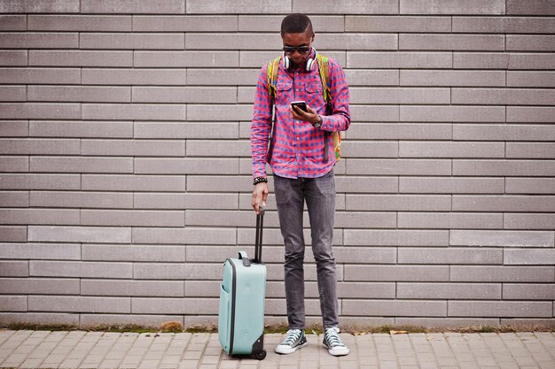 African american man in checkered shirt sunglasses and earphones with suitcase and backpack Black man traveler against wall holding mobile phone